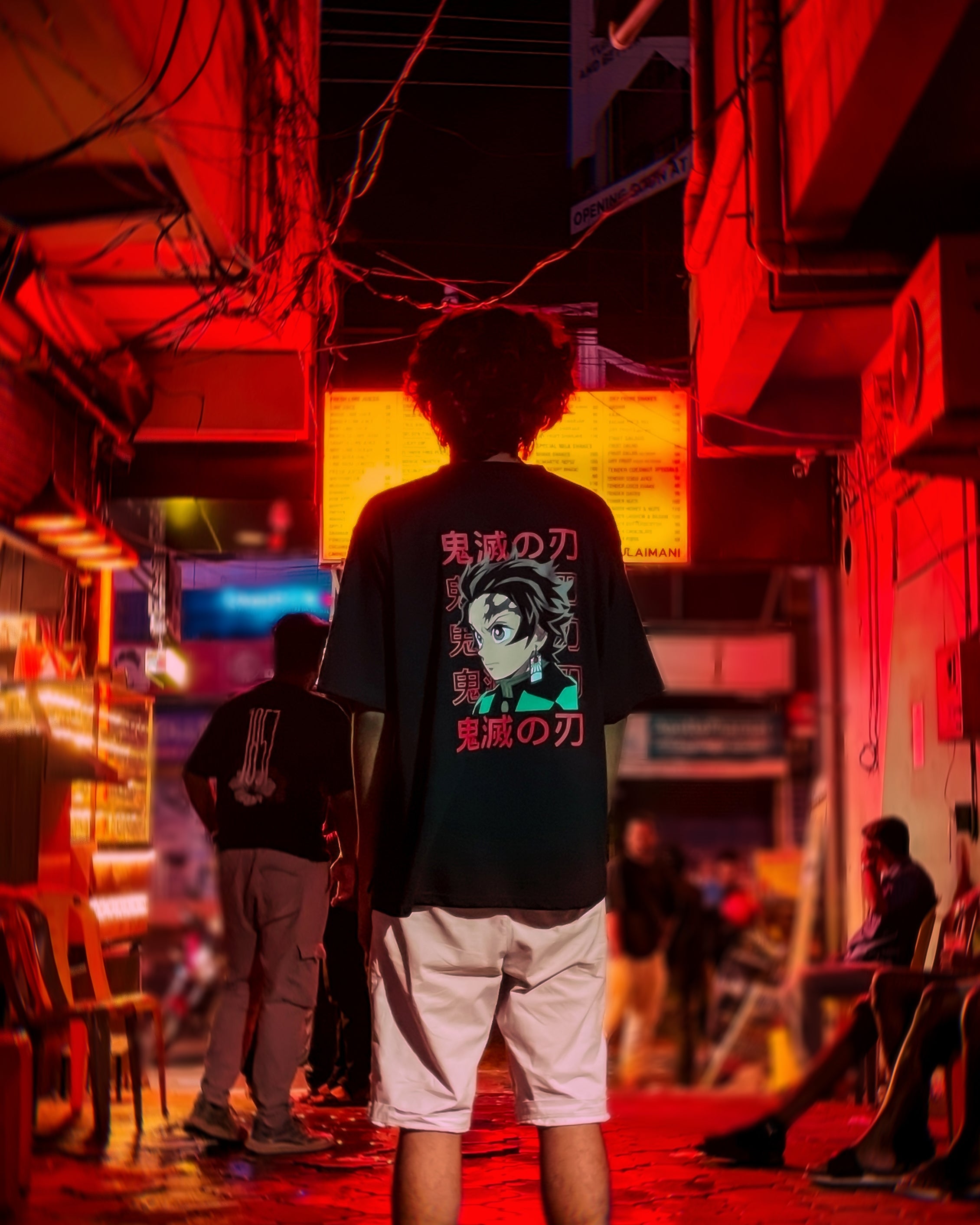 A person with curly hair, wearing the Tanjiro Kamado Oversized Tshirt and white shorts, stands in a neon-lit alley at night. Red lights illuminate the scene with blurred figures and signs in the background.
