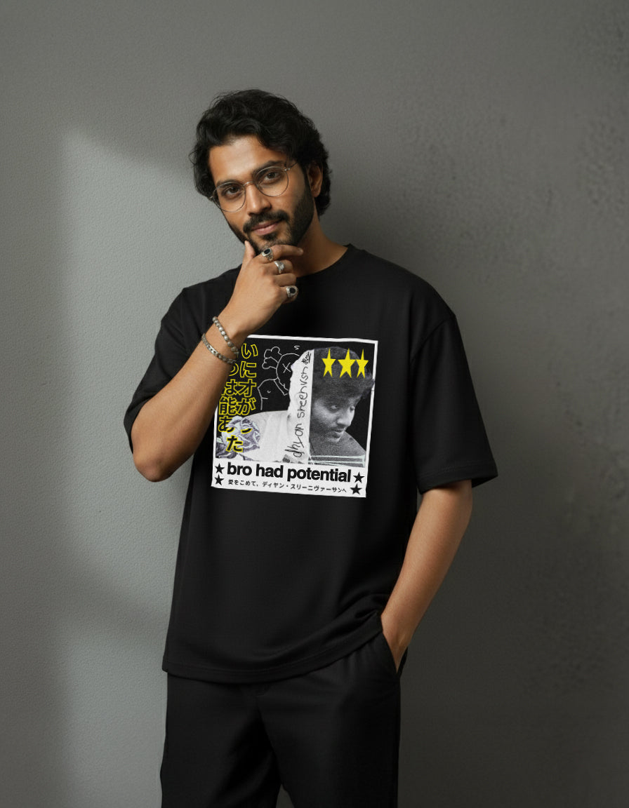A man with glasses and a beard stands against a gray textured wall, wearing an oversized black Japan Dhyan 220 GSM Heavy Weight T-shirt featuring graphic designs, yellow text, three yellow stars, and the phrase “bro had potential,” hand on chin.