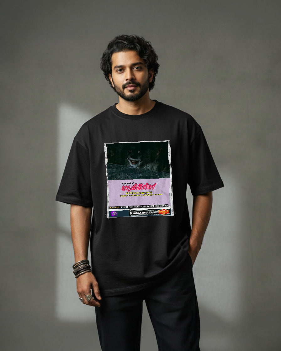 A man with wavy hair and a beard stands against a gray background, wearing the Oodayil Ninnu IT oversized t-shirt made from 220 GSM cotton with a colorful chest graphic. He looks neutral, one hand in his pocket.