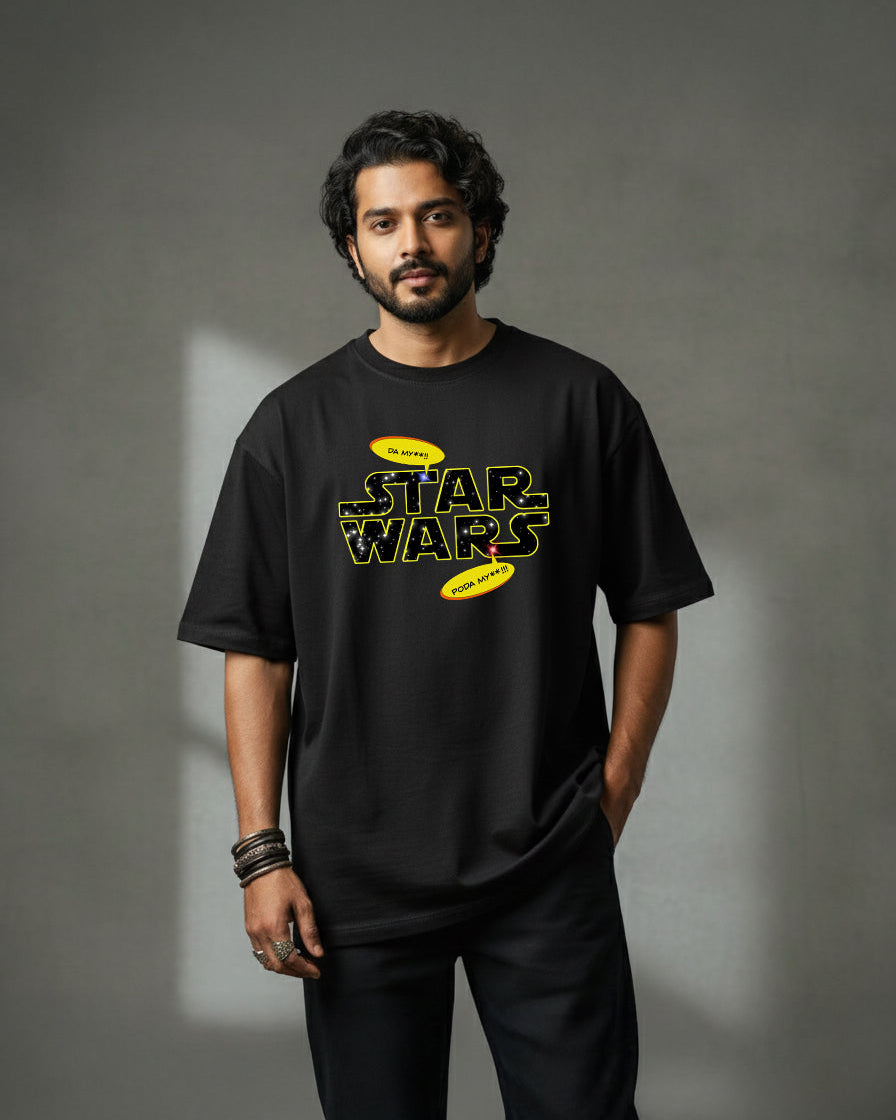 A man with dark hair and a beard stands against a grey background, wearing a loose black bio-washed Star Wars t-shirt featuring yellow lettering and black pants. One hand is in his pocket, and he wears several bracelets.